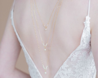 May include: A layered back necklace with small pearls on a gold chain. The necklace is worn by a model with a white lace dress.
