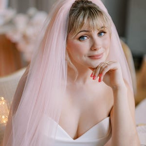 May include: A bride wearing a white strapless wedding dress and a pink veil. The veil is draped over the head and shoulders. The bride has fair skin, blue eyes, and is wearing a ring on her finger.