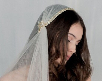 Puede incluir: Un velo de tul blanco transparente con una diadema dorada adornada con intrincados detalles metálicos y pequeños cristales transparentes. El velo está sujeto al lateral de la diadema con un adorno en forma de flor.