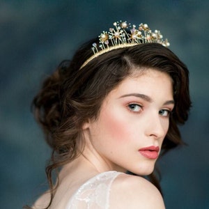 May include: A gold bridal crown with delicate floral details and iridescent crystals. The crown is worn by a woman with brown hair and makeup.