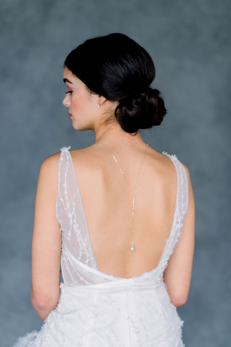 May include: A white bridal gown with a low back and a delicate pearl necklace. The necklace is a long, thin chain with a single pearl pendant.