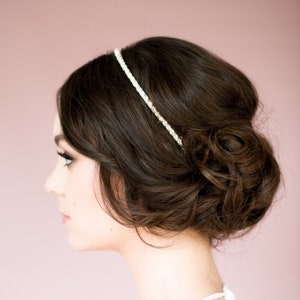 May include: A woman's dark brown hair styled in an updo, adorned with a delicate, sparkling headband. The headband is thin and features small, glittering embellishments. The background is a soft, muted pink.