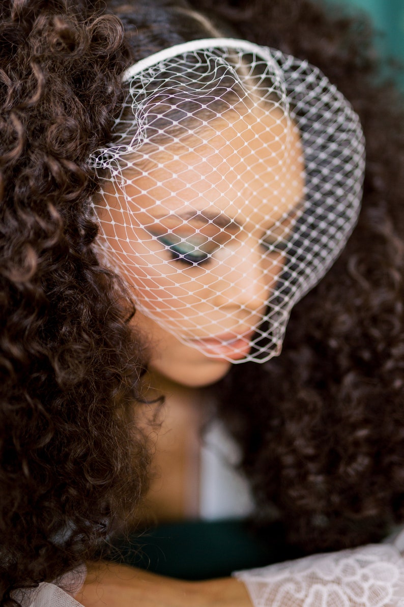 May include: A woman with curly brown hair wears a white veil over her face. The veil is a fine mesh with a delicate pattern. The woman's eyes are closed and her lips are slightly parted.