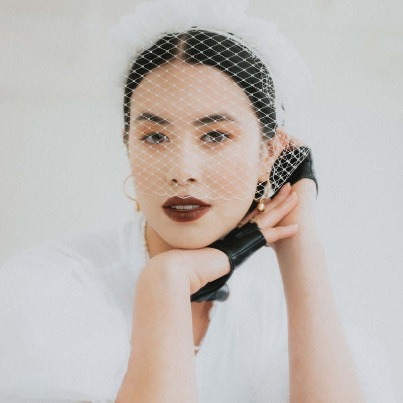 May include: A woman with dark hair is wearing a white veil and black leather gloves. She is looking at the camera with a serious expression. Her lips are painted a deep red color.