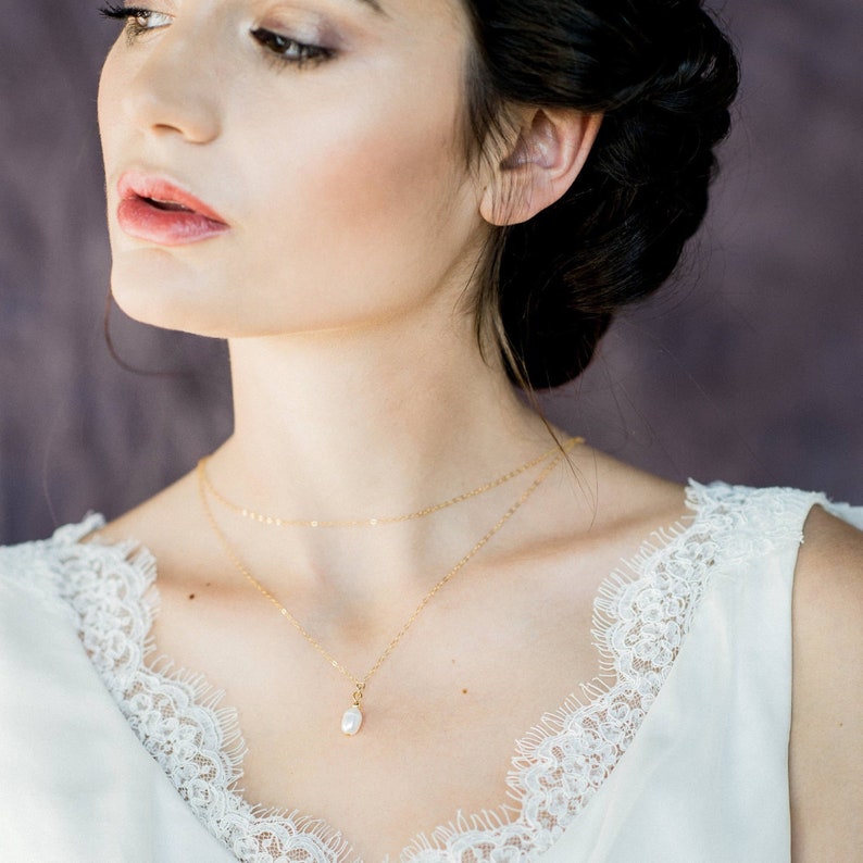 May include: A woman wearing a white lace top with a delicate gold necklace featuring a single pearl pendant.