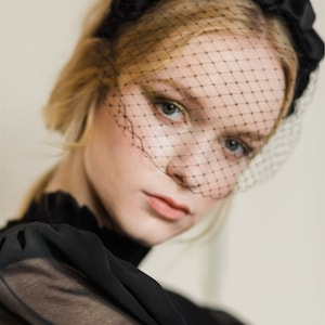 May include: A black satin headband with a gathered detail and a black mesh veil. The veil covers the forehead and eyes.