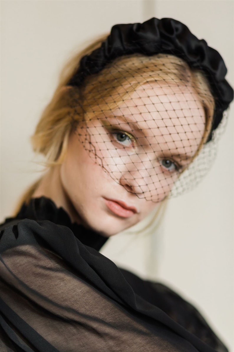 May include: A black satin headband with a gathered detail and a black mesh veil. The veil covers the forehead and eyes.