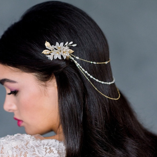 May include: A gold and white floral hair comb with a delicate chain attached. The comb is worn in a woman's dark hair.