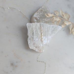 May include: A silver chain necklace with a single pearl pendant. The necklace is layered with a shorter chain with small pearls. The necklace is displayed on a white surface with a piece of selenite.