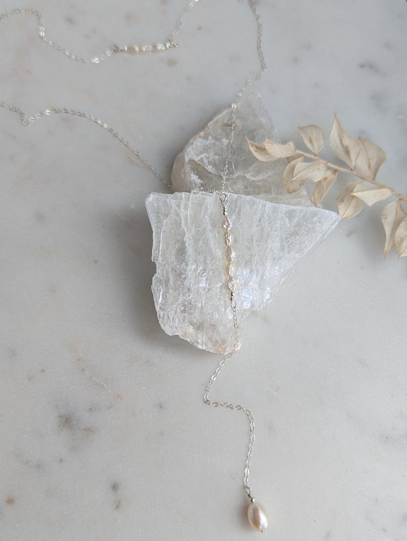 May include: A silver chain necklace with a single pearl pendant. The necklace is layered with a shorter chain with small pearls. The necklace is displayed on a white surface with a piece of selenite.