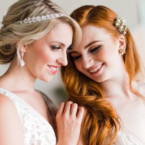 May include: Two people wearing white lace dresses, one with a sparkling headband and the other with a gold and pearl hair clip. They are smiling and looking at each other.