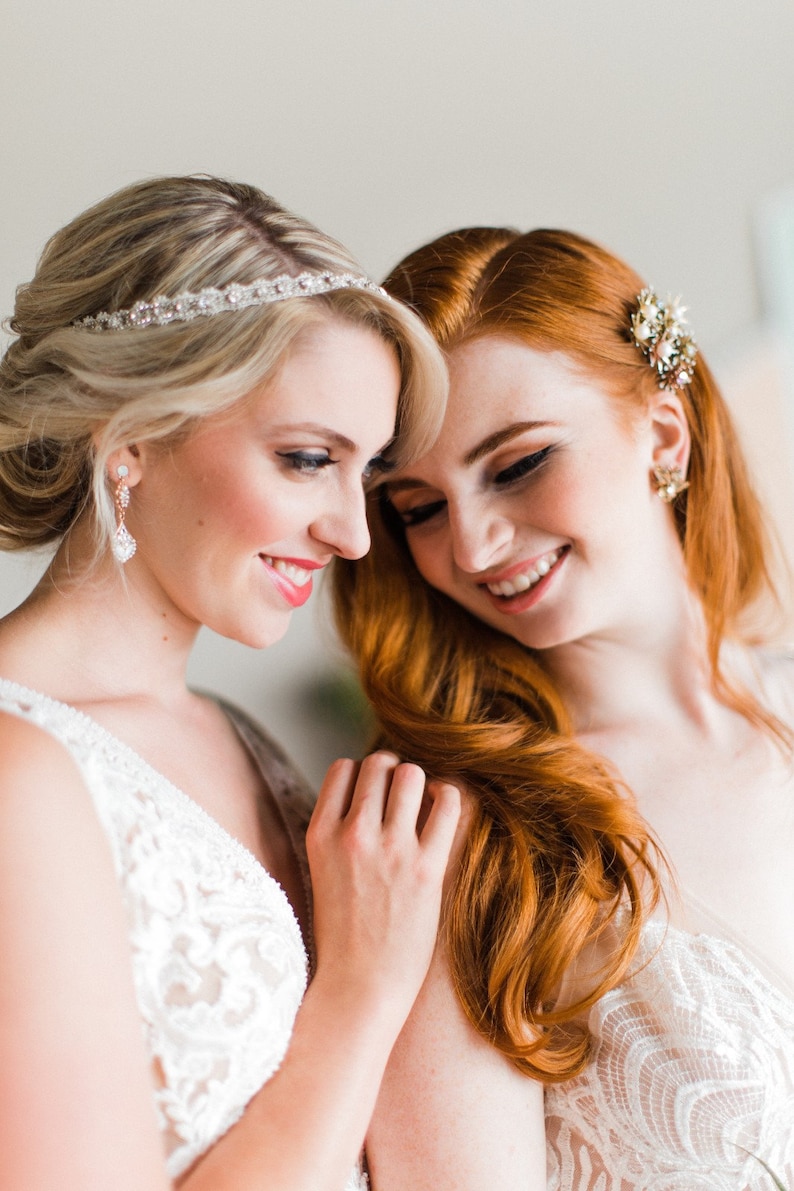 May include: Two people wearing white lace dresses, one with a sparkling headband and the other with a gold and pearl hair clip. They are smiling and looking at each other.