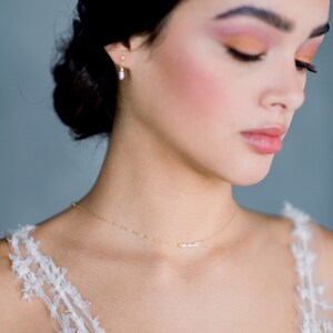 May include: A woman wearing a white lace top with a delicate gold necklace featuring small pearls. The necklace is a simple and elegant piece of jewelry.