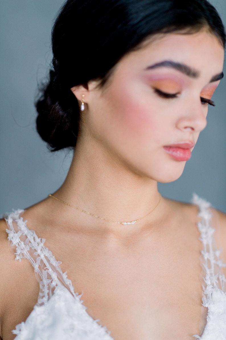 May include: A woman wearing a white lace top with a delicate gold necklace featuring small pearls. The necklace is a simple and elegant piece of jewelry.