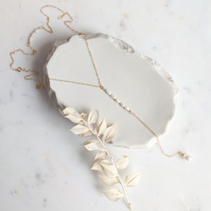 May include: A delicate gold chain necklace with a single strand of pearls hanging from the center. The necklace is displayed on a white marble surface with a dried branch.