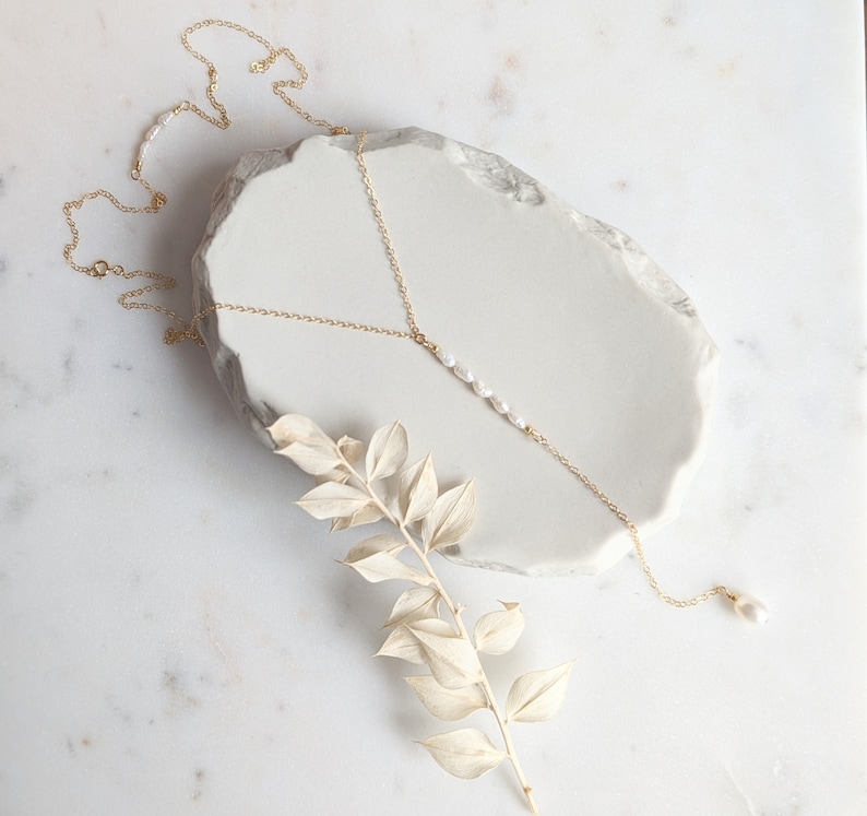May include: A delicate gold chain necklace with a single strand of pearls hanging from the center. The necklace is displayed on a white marble surface with a dried branch.
