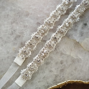 May include: Two white satin ribbon garters with a silver rhinestone embellishment. The embellishment is a floral design with a beaded center.