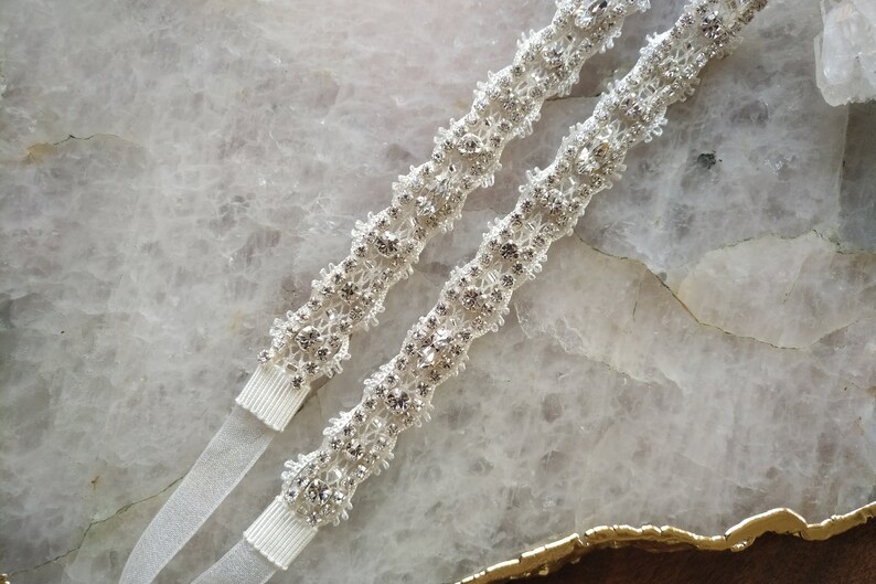 May include: Two white satin ribbon garters with a silver rhinestone embellishment. The embellishment is a floral design with a beaded center.