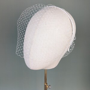 May include: A white birdcage veil with a delicate net pattern. The veil is on a white mannequin head and is ready to be worn for a wedding or special occasion.