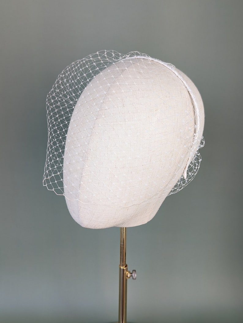 May include: A white birdcage veil with a delicate net pattern. The veil is on a white mannequin head and is ready to be worn for a wedding or special occasion.