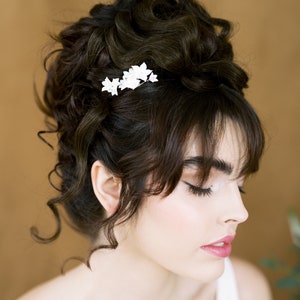 Può includere: Una donna con i capelli castano scuro raccolti in un alto chignon con fermagli per capelli a forma di fiori bianchi. Indossa una canotta bianca senza maniche e ha gli occhi chiusi.