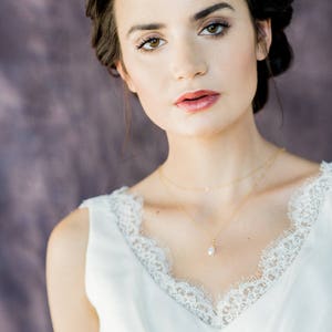 May include: A woman with dark hair and a white lace top is wearing a delicate gold necklace with a pearl pendant.