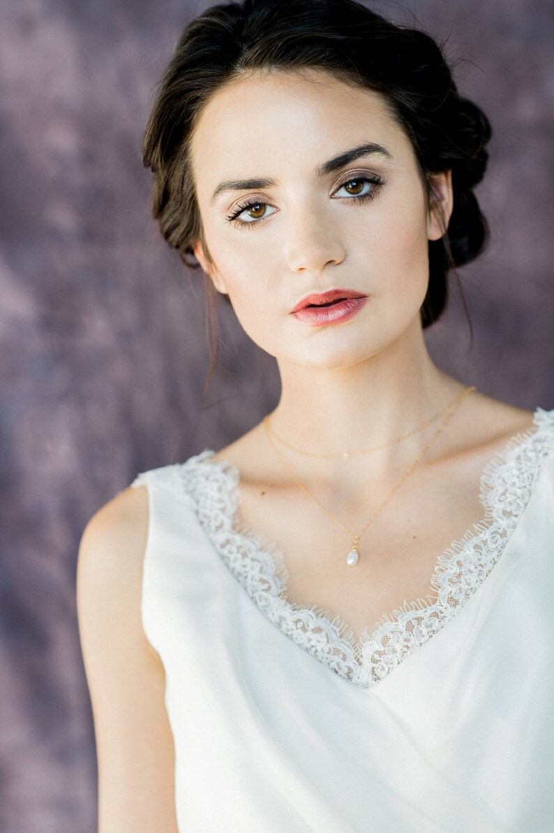 May include: A woman with dark hair and a white lace top is wearing a delicate gold necklace with a pearl pendant.