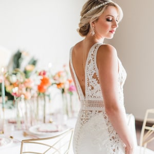 May include: A bride wearing a white lace wedding dress with a low back and a beaded headband. The dress has a fitted bodice and a flowing skirt. The bride is standing in front of a table with flowers and candles.