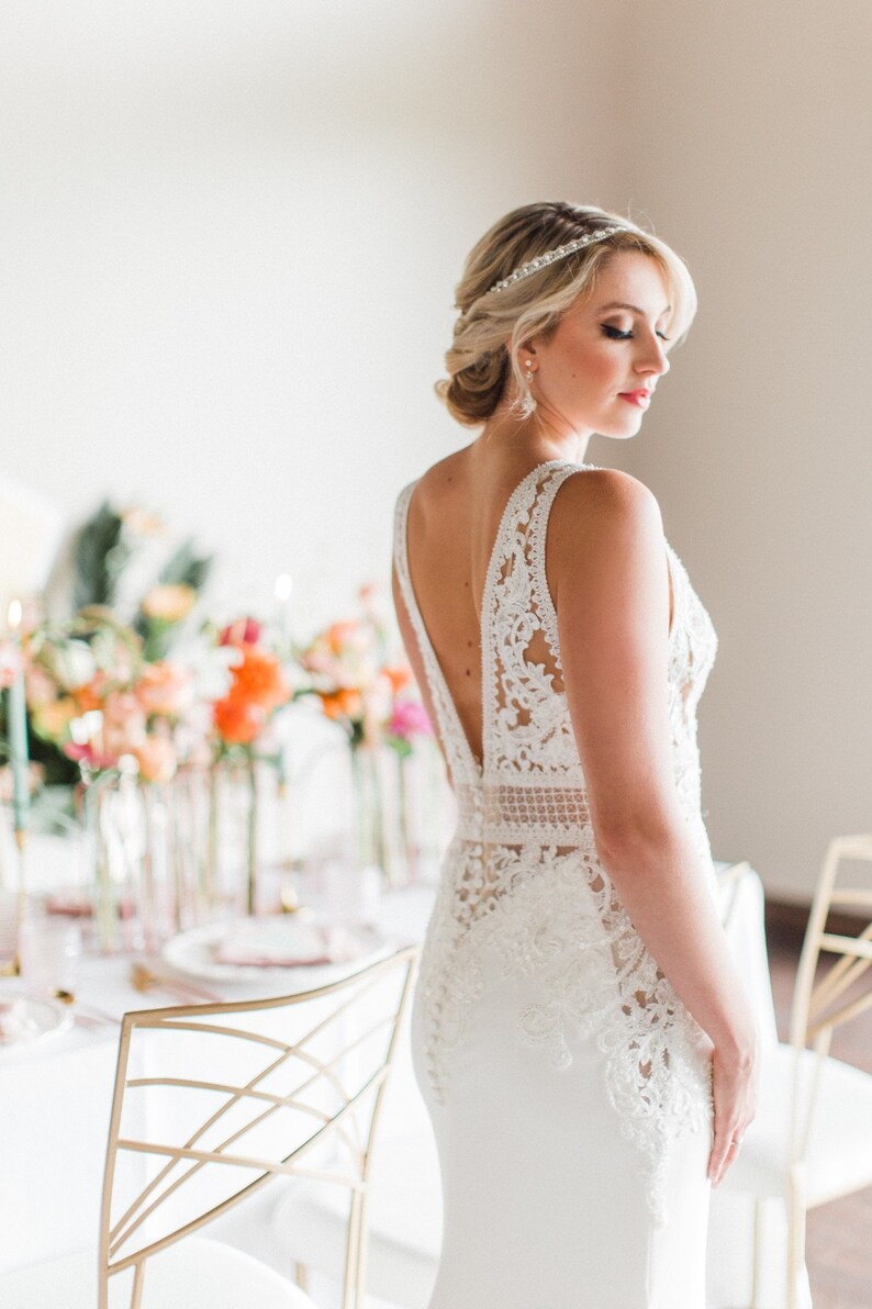 May include: A bride wearing a white lace wedding dress with a low back and a beaded headband. The dress has a fitted bodice and a flowing skirt. The bride is standing in front of a table with flowers and candles.