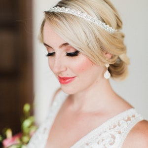 May include: A bride with blonde hair wearing a white lace wedding dress and a sparkly headband. She is wearing delicate earrings and has her hair pulled back.