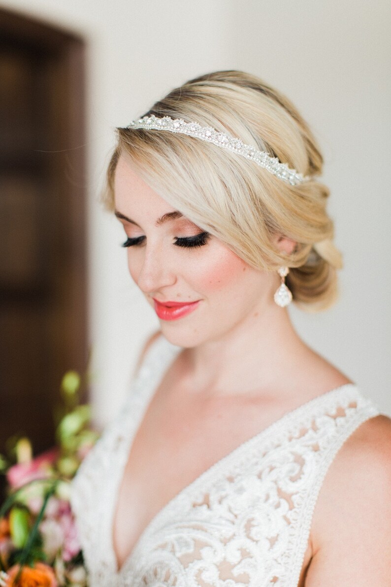 May include: A bride with blonde hair wearing a white lace wedding dress and a sparkly headband. She is wearing delicate earrings and has her hair pulled back.