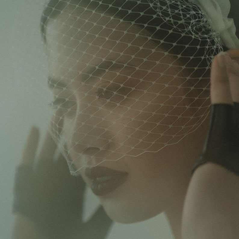 May include: A woman wearing a white veil with a fine mesh pattern. The veil covers her face and hair. She is wearing dark lipstick and has a serious expression.