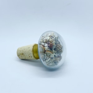 May include: A clear glass bottle stopper with a cork base filled with small seashells. The seashells are a mix of white, brown, and tan.