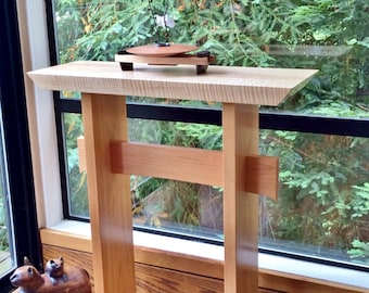 May include: A wooden table with a natural wood finish and a simple design. The table has a rectangular top and two legs on each side. The table is in front of a window with a view of trees.