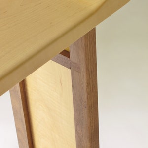 May include: Close-up of a wooden table with a light wood top and dark wood legs. The table top is slightly rounded and the legs are joined together with a mortise and tenon joint.