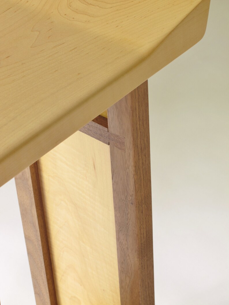 May include: Close-up of a wooden table with a light wood top and dark wood legs. The table top is slightly rounded and the legs are joined together with a mortise and tenon joint.