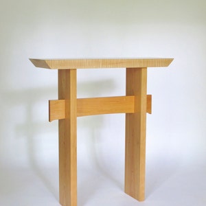 May include: A wooden console table with a light wood finish. The table has a simple design with a rectangular top and two vertical supports. The supports are connected by a horizontal bar.