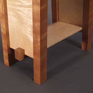 May include: A wooden end table with a single shelf. The table is made of light wood with a darker wood trim. The table has a square base.
