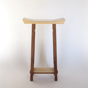 May include: A wooden stool with a light wood top and dark wood legs. The stool has a curved top and a shelf underneath.