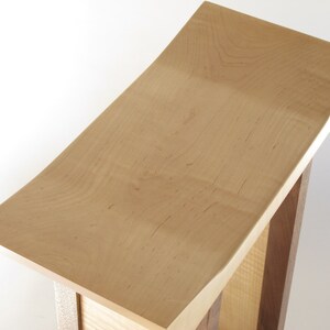 May include: A wooden stool with a curved, light brown top and a base made of darker wood.