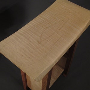 May include: A small wooden end table with a curved top made of maple wood. The table has a single shelf and is supported by four legs.
