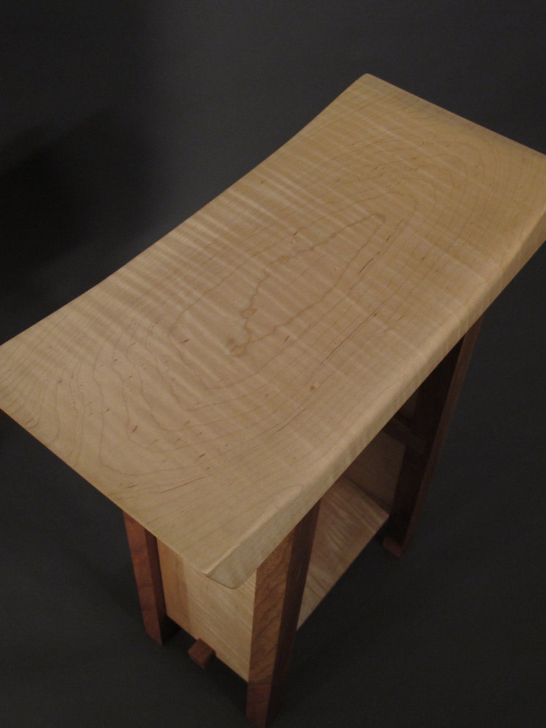 May include: A small wooden end table with a curved top made of maple wood. The table has a single shelf and is supported by four legs.