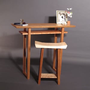 May include: A wooden table with a shelf and a matching stool. The table has a light wood finish and the stool has a natural wood finish. There is a framed photograph and a vase with flowers on the table.