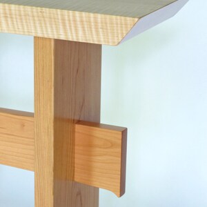 May include: A light wood console table with a simple, modern design. The table has a rectangular top with a beveled edge and a single, square leg with a cross brace.