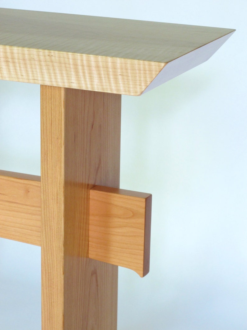 May include: A light wood console table with a simple, modern design. The table has a rectangular top with a beveled edge and a single, square leg with a cross brace.