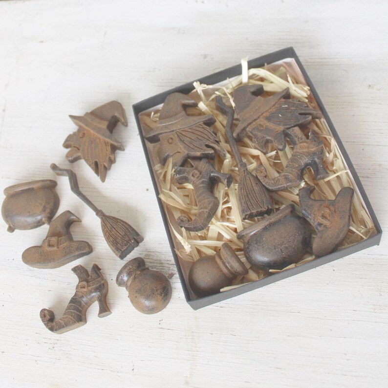 Op de afbeelding: Een set van 10 Halloween-figuurtjes gemaakt van donkerbruin metaal. De figuurtjes omvatten heksen, bezems, ketels en hoeden. De figuurtjes zijn gerangschikt in een doos met bruin, versnipperd papier.