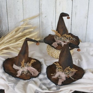 May include: Three brown witch hats with frayed edges and brown accents. Each hat has a small brown acorn on the tip and is decorated with dried grasses and a piece of brown burlap.