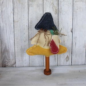 May include: A whimsical, handmade tree-shaped decoration. It features a navy blue fabric hat, a beige body, and a yellow base. A small mitten with red accents and sprigs of greenery are attached. The tree is mounted on a wooden dowel and base.