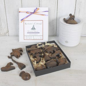 Op de afbeelding: Een set Halloween-thema wax melts in een zwarte doos met een bruine papieren inleg. De wax melts zijn gevormd als heksen, bezems, ketels en hoeden. De doos is vastgebonden met een paars en oranje lint. De tekst op de doos luidt "Kenzie's Cottage Witches Brew Handmade Wax Melts".