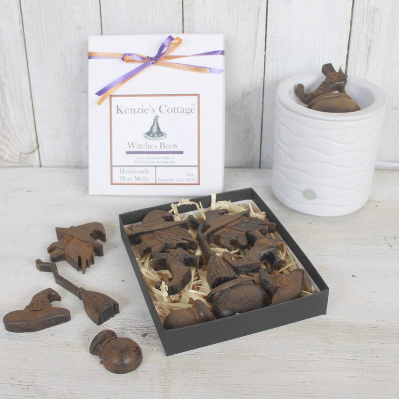 Op de afbeelding: Een set Halloween-thema wax melts in een zwarte doos met een bruine papieren inleg. De wax melts zijn gevormd als heksen, bezems, ketels en hoeden. De doos is vastgebonden met een paars en oranje lint. De tekst op de doos luidt "Kenzie's Cottage Witches Brew Handmade Wax Melts".